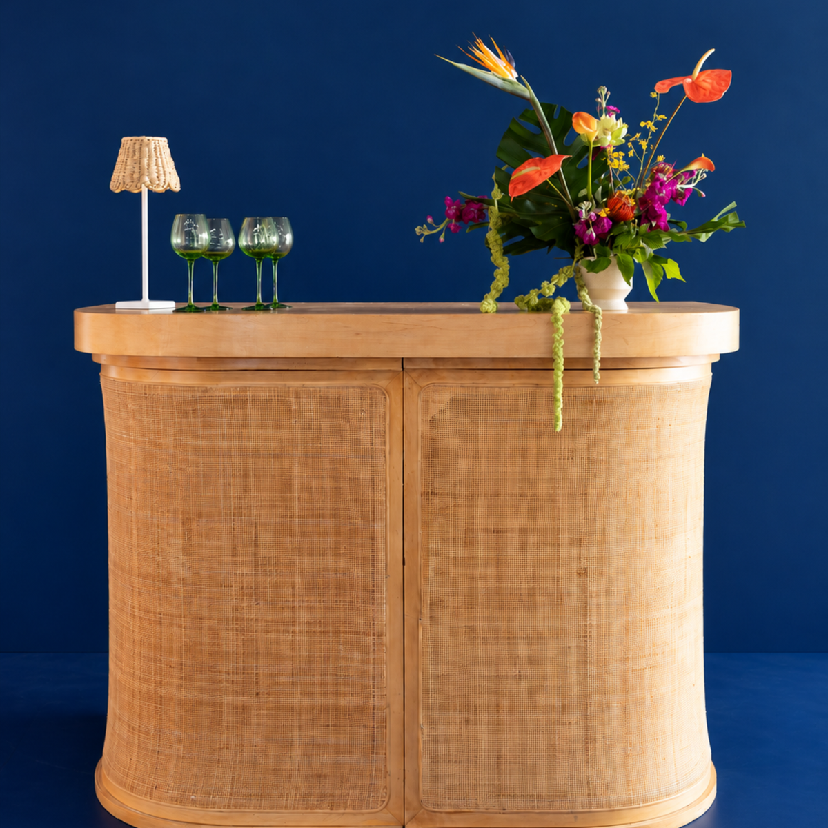 A curved wooden console table with woven cane panels, styled with a small lamp, two green wine glasses, and a vibrant flower arrangement against a dark blue background.