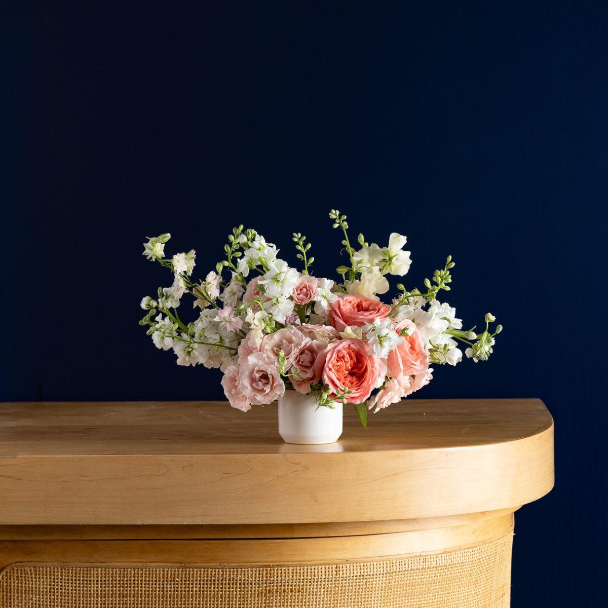A floral arrangement of pink and white flowers in a small white vase, placed on a light wood console table against a dark navy blue background.