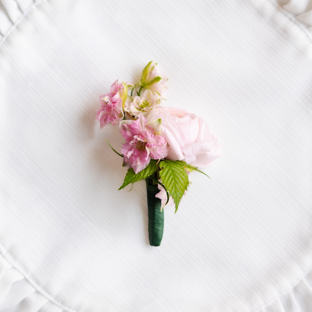 A small floral boutonniere featuring light pink flowers and green leaves, wrapped in a dark green ribbon and placed on a white textured fabric background.