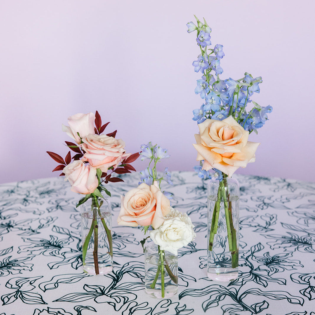 Three clear glass bud vases of varying sizes, arranged on a table with a striped orange and white tablecloth, containing floral arrangements.