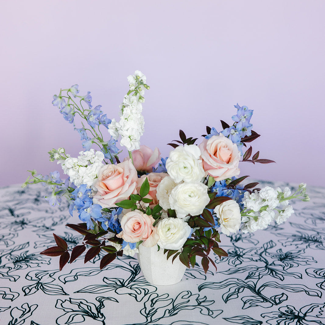 A colorful floral centerpiece arranged in a white ceramic vase, featuring a variety of flowers in orange, purple, and pink shades, placed on a table with a striped orange and white tablecloth.