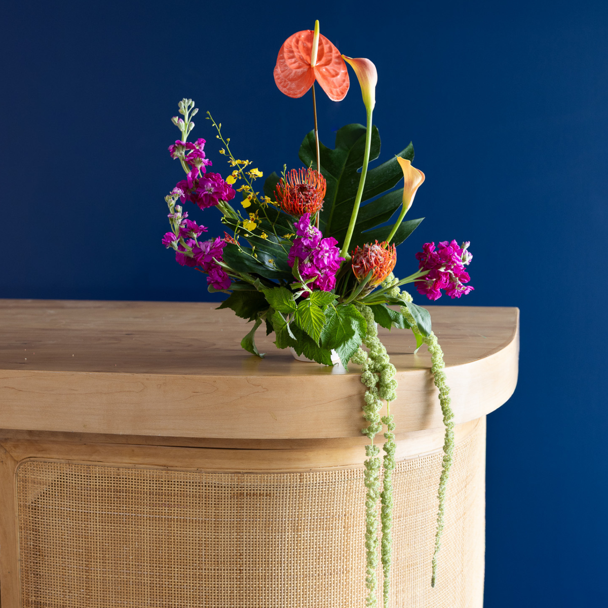 A vibrant floral arrangement featuring a coral anthurium, magenta flowers, green foliage, and trailing greenery, displayed on a light wood console table against a deep blue wall.