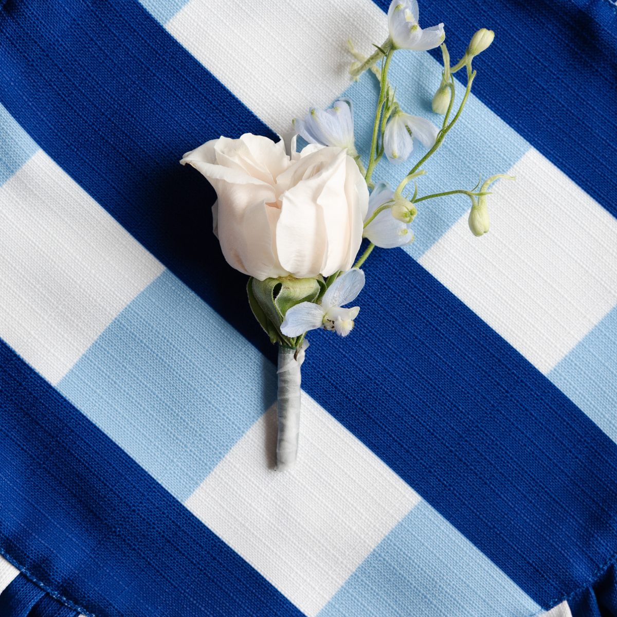 A floral boutonniere with a pale blush rose and small light blue flowers, arranged on a blue and white striped fabric background.