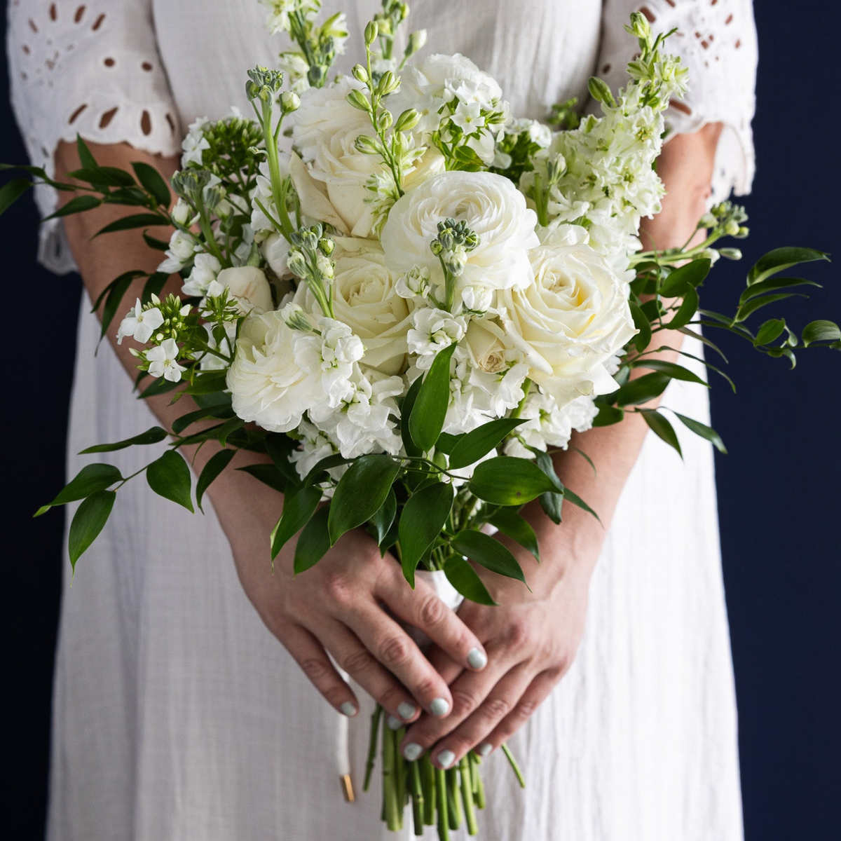 A close-up of a person in a white dress holding a large white floral bouquet with white roses, white snapdragons, and green foliage against a dark navy background.