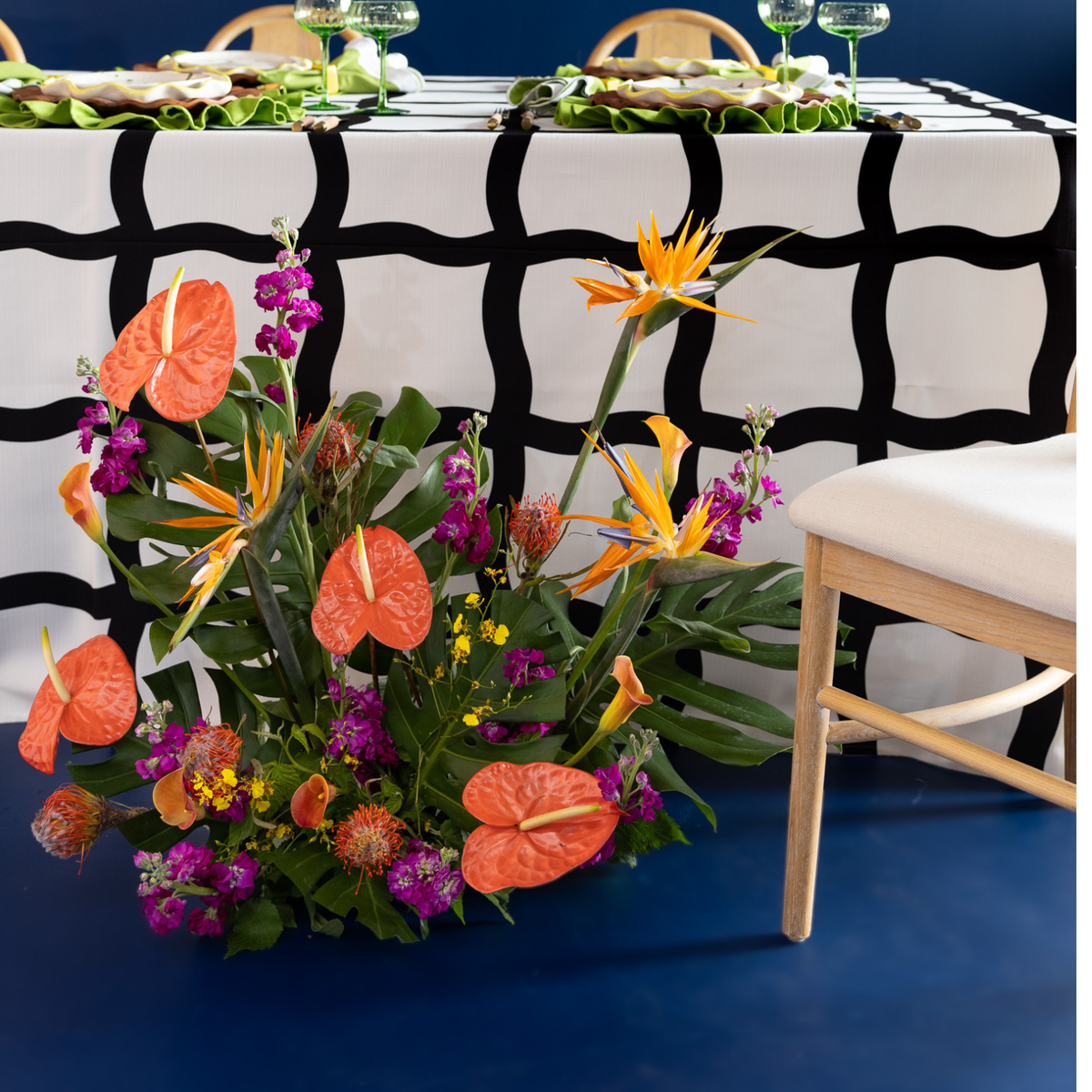 A vibrant floral arrangement featuring orange anthuriums, bird of paradise flowers, and purple blossoms with green foliage, styled on a dark blue floor in front of a table with a black and white patterned tablecloth.