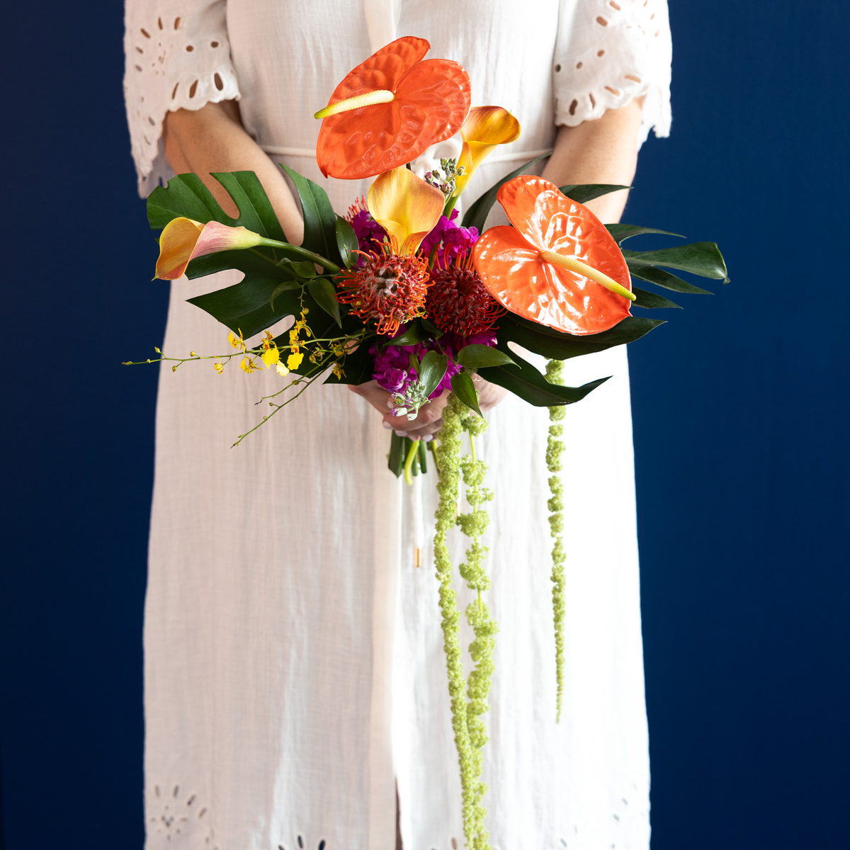 A person in a white eyelet dress holds a vibrant tropical flower bouquet featuring orange anthuriums, yellow calla lilies, green monstera leaves, and hanging green bead vines against a dark blue background.