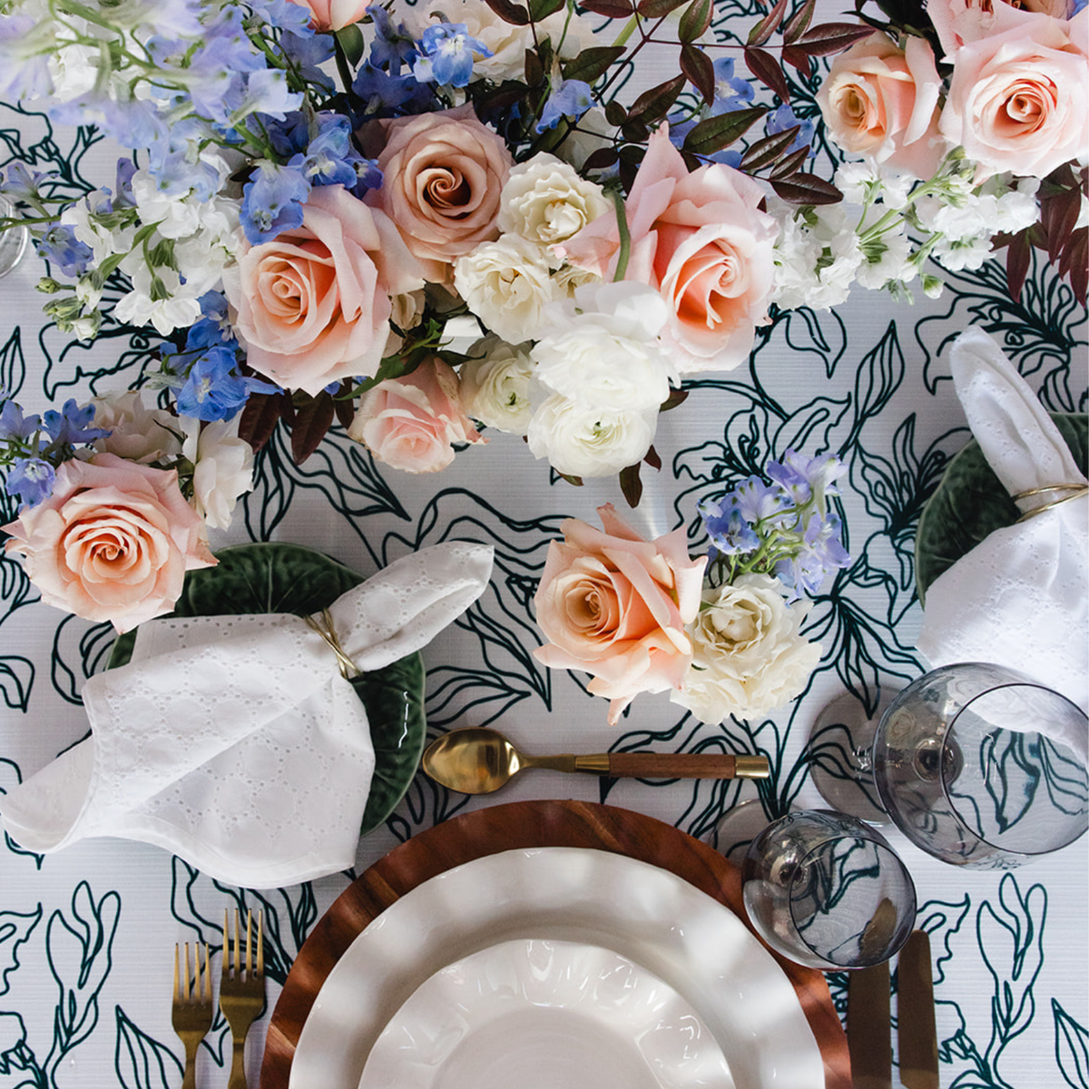 An overhead styled table setting featuring a floral centerpiece with blush pink, white, and blue flowers on a white tablecloth with a dark green botanical pattern. The setting includes white plates with a copper rim, white napkins tied with gold rings, gold flatware, and clear drinking glasses.