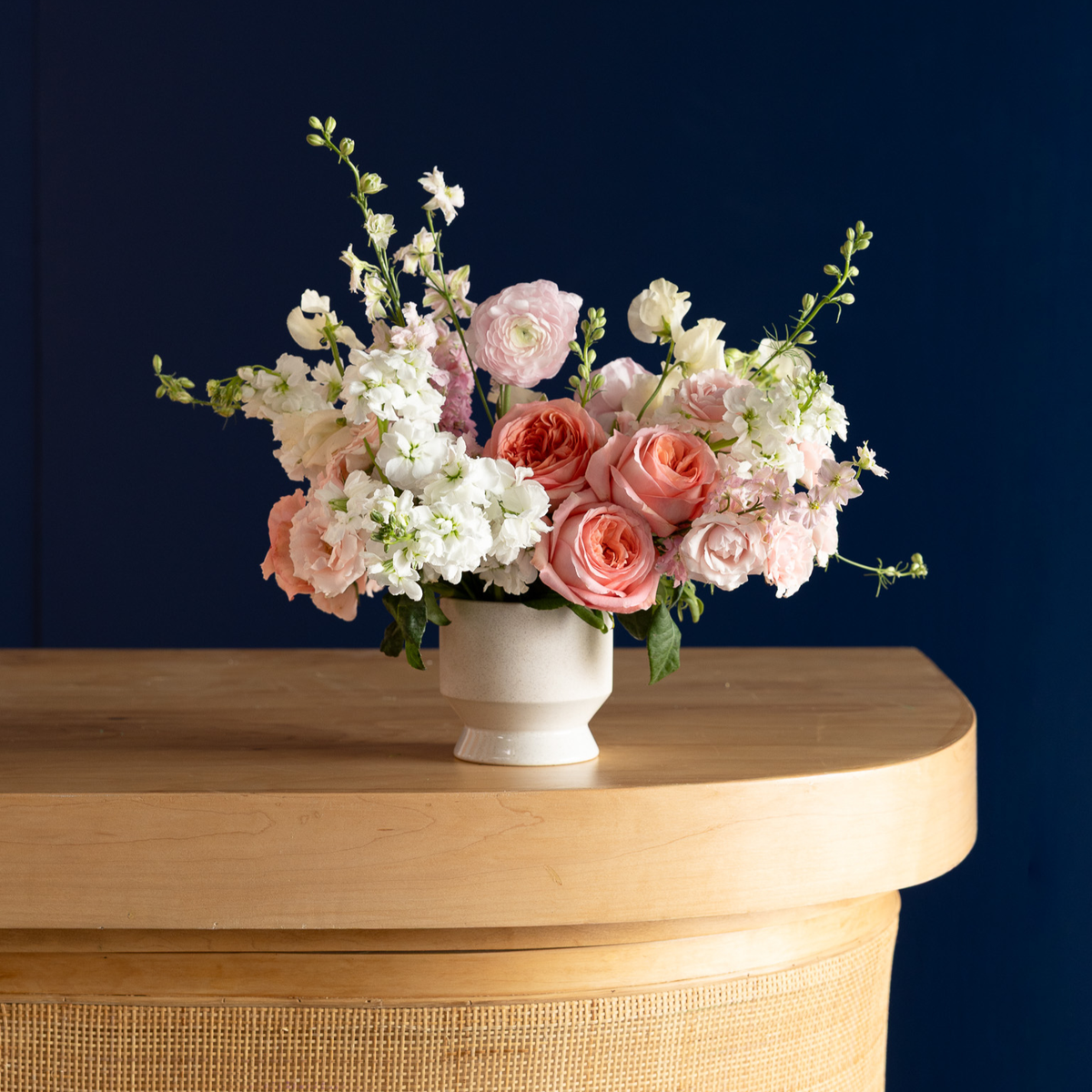 A floral arrangement of pink and white roses, hydrangeas, and snapdragons in a small white ceramic pot, displayed on a light wood console table against a dark navy blue wall.