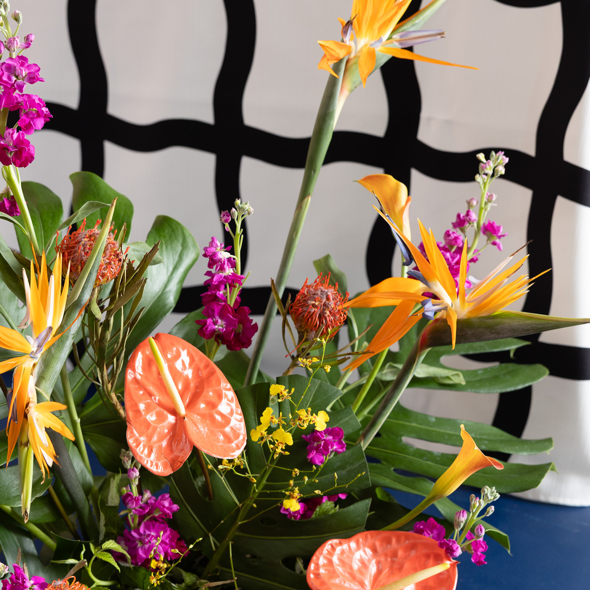 A vibrant tropical flower arrangement featuring orange anthuriums, yellow bird of paradise, bright pink spikes, green monstera and philodendron leaves, and small yellow blossoms, set against a black and white wavy patterned backdrop.