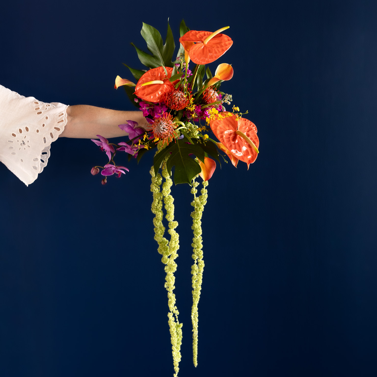 A vibrant floral bouquet held by a person's arm, featuring bright orange anthuriums, green leaves, purple flowers, and long hanging lime-green seed pods, set against a solid dark navy blue background.