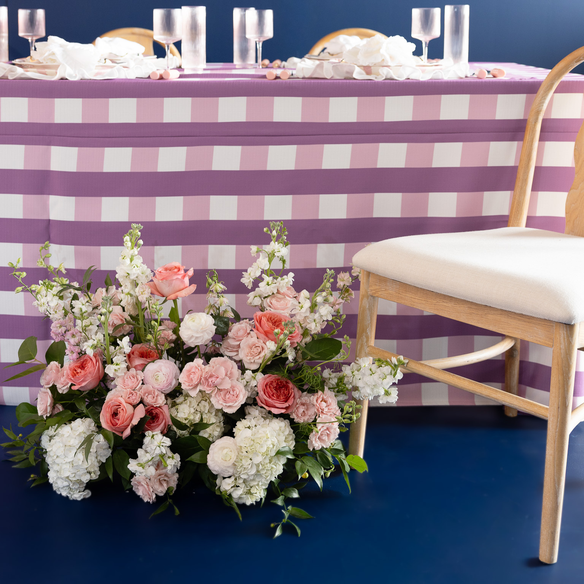 A pink and white checkered tablecloth draped over a long table, styled with white napkins, wine glasses, and a large pink and white floral arrangement on a dark blue floor next to a wooden chair with a cream seat.