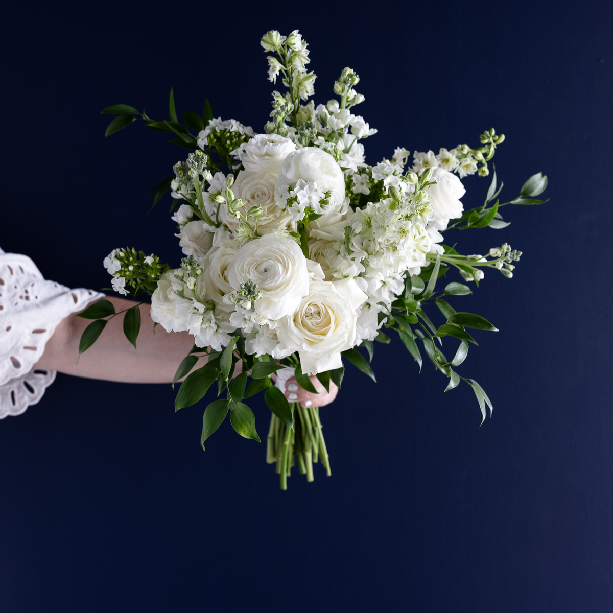A hand holding a white floral bouquet with roses, ranunculus, and green foliage against a dark navy blue background.