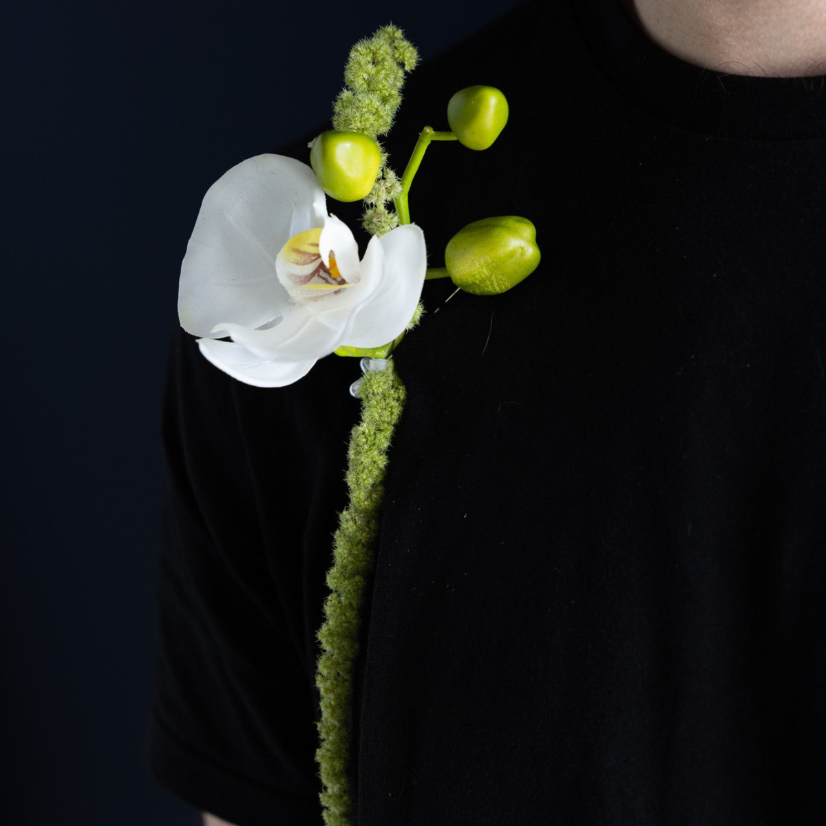A white orchid flower with green buds attached to a person's black shirt against a dark navy background.