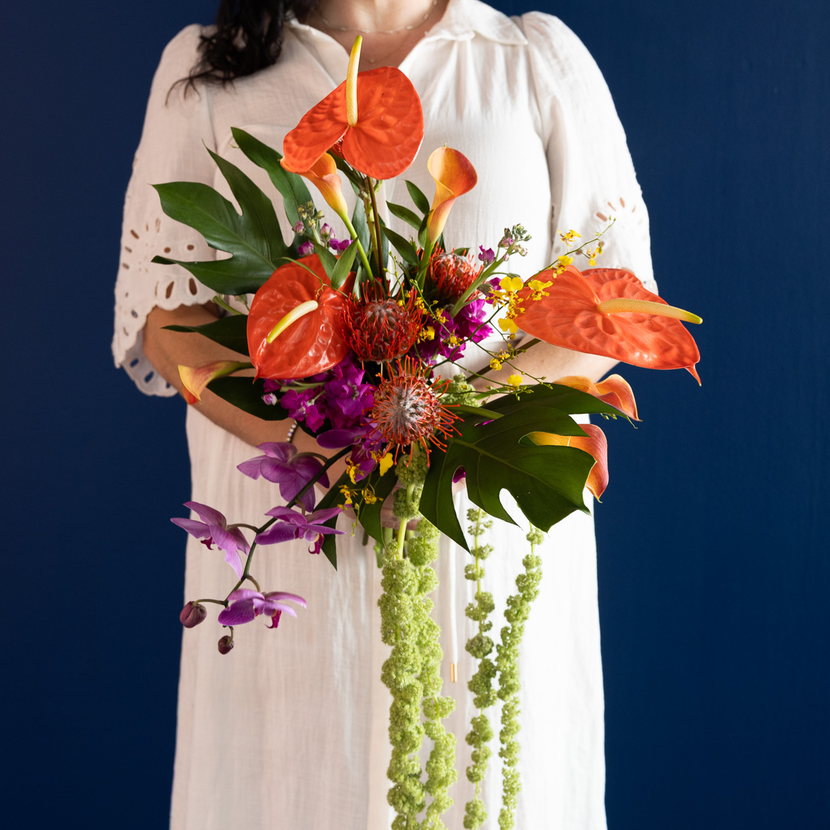 A person in a white dress holds a vibrant tropical flower bouquet featuring orange anthuriums, purple orchids, green trailing foliage, and red protea against a dark blue background.