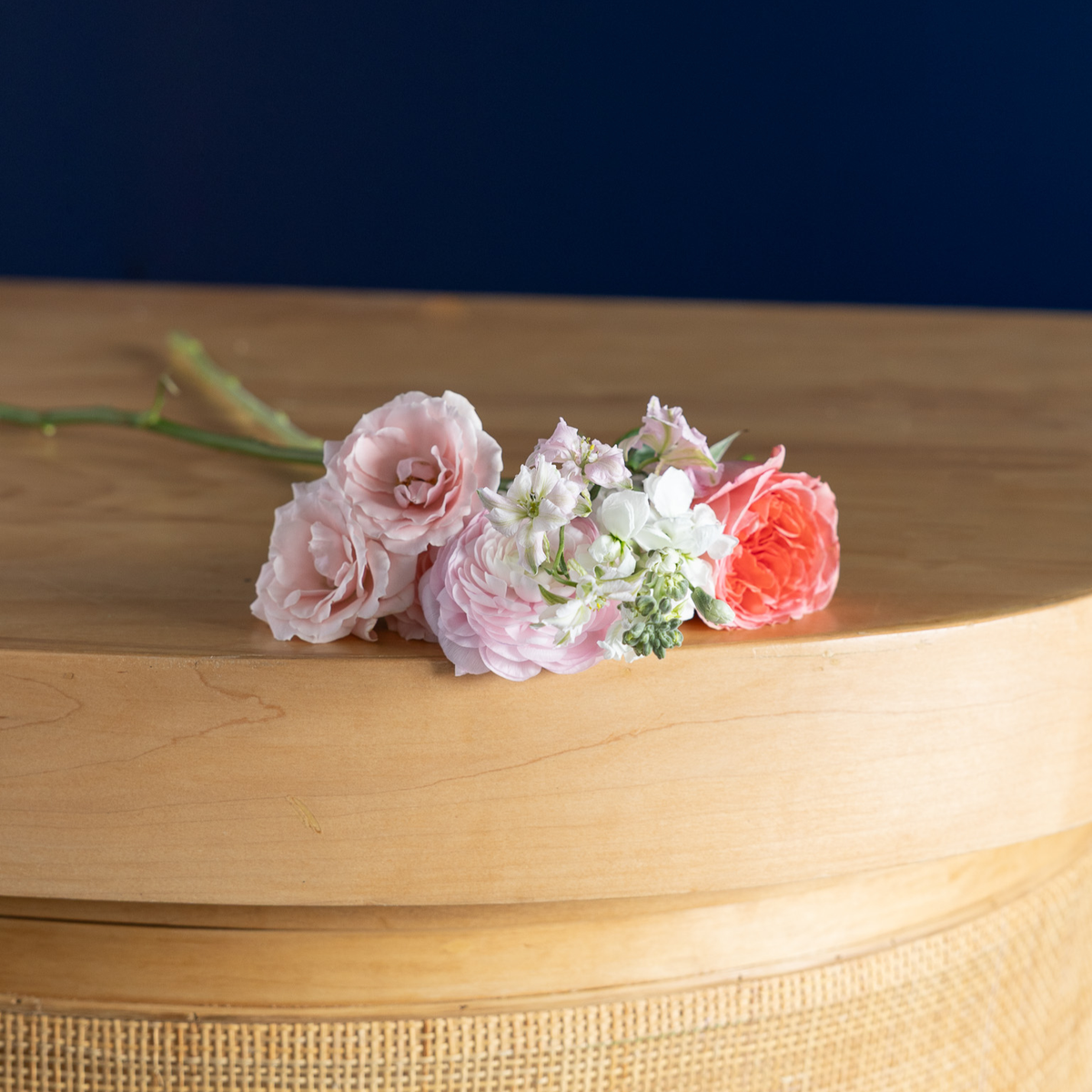 A small floral arrangement of pink and coral roses with white filler flowers and green stems, placed on a curved light wood surface against a dark navy background.
