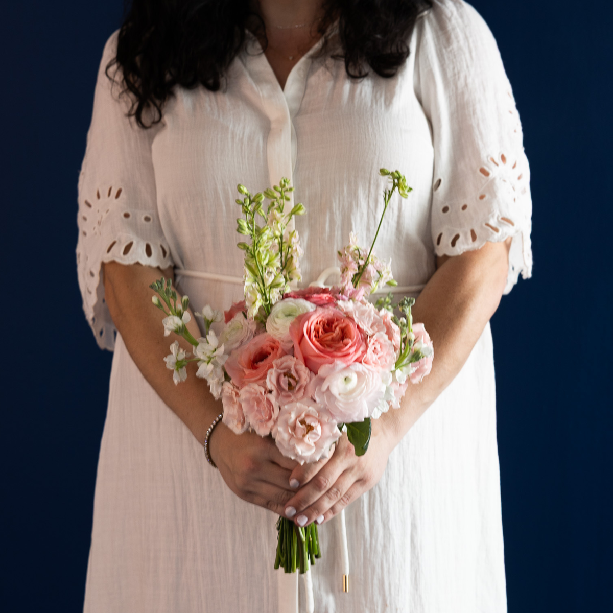 A woman in a white eyelet dress holds a pink and white floral bouquet against a dark navy blue background.