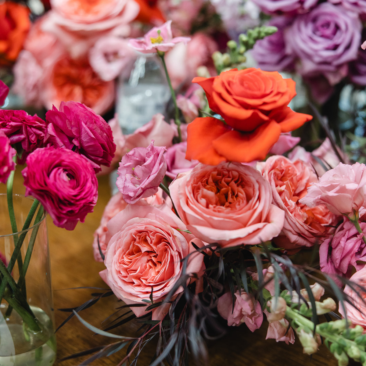 A close-up floral arrangement featuring vibrant pink, coral, and orange roses with dark burgundy filler stems, displayed in a glass vase on a wooden surface.
