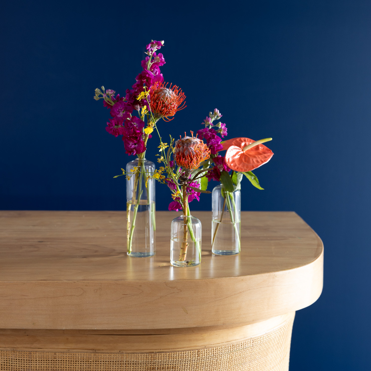 Three clear glass vases filled with colorful artificial flowers, including magenta blossoms, orange protea-like blooms, and a red anthurium, arranged on a light wood table against a dark blue background.