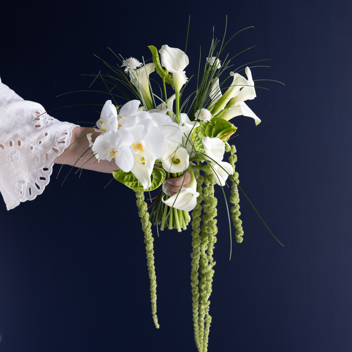 A hand holding a white floral bouquet with calla lilies, white orchids, green foliage, and long hanging green stems against a dark navy background.