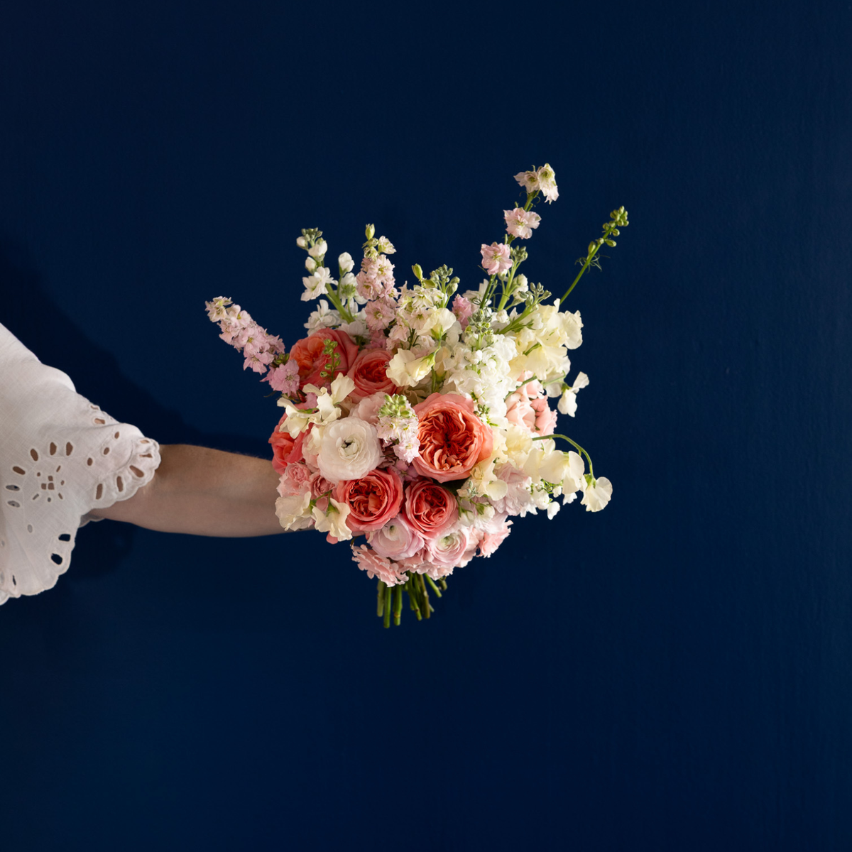 A hand holding a pastel floral bouquet with pink and white flowers against a dark navy blue background.
