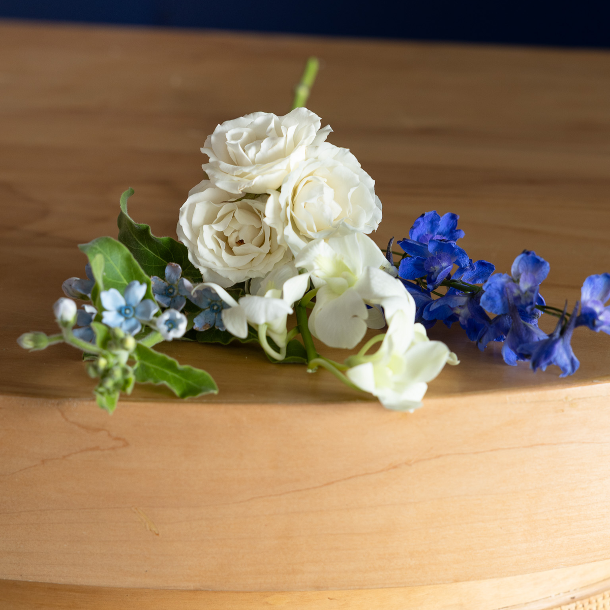 A small floral arrangement with white roses, blue delphiniums, and green leaves resting on a light wood surface.