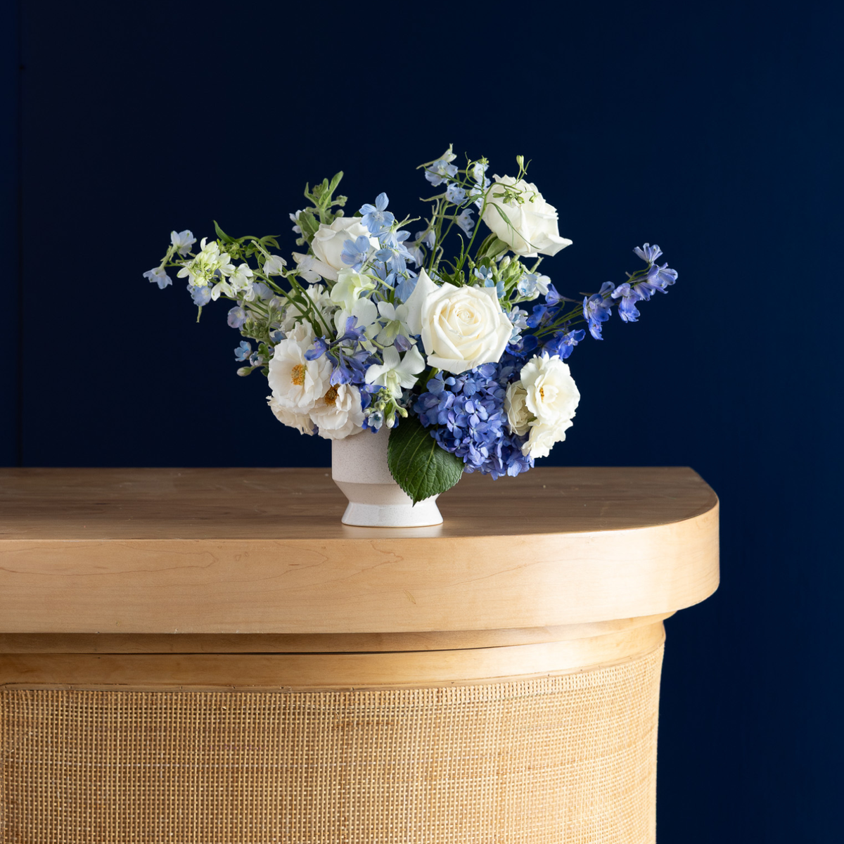 A floral arrangement featuring white roses, blue hydrangeas, and blue delphiniums in a small white ceramic pot, displayed on a light wood console table against a dark navy blue background.