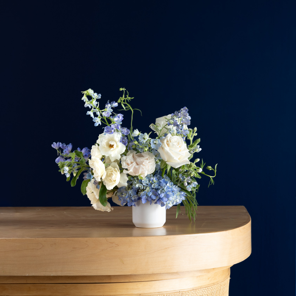 A floral arrangement of white and blue flowers in a small white vase, placed on a light wood console table against a dark navy background.