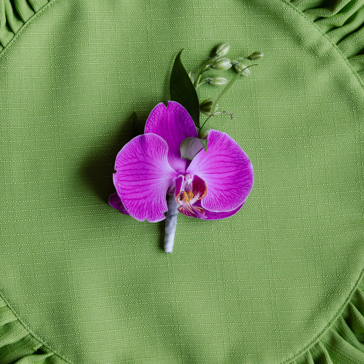A single purple orchid flower boutonniere with green leaves and buds, arranged on a textured olive green fabric surface.