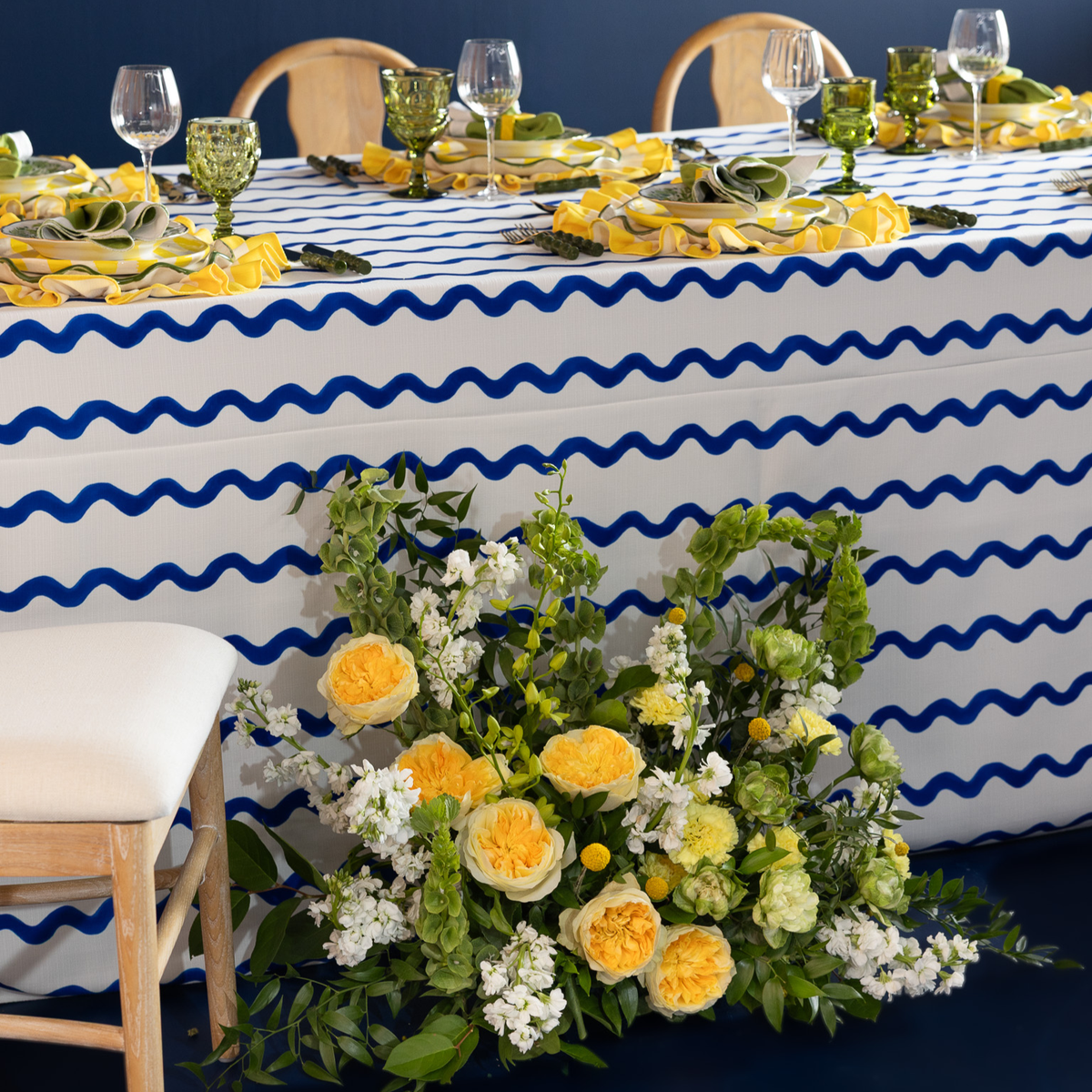 A styled event table setup featuring a white tablecloth with a bold blue wavy stripe pattern, yellow and green tableware, floral centerpieces, and wooden chairs against a dark blue wall.