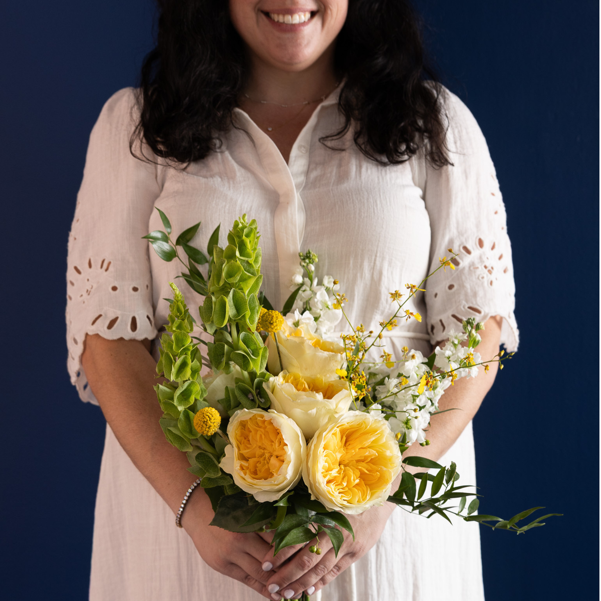 A woman in a white eyelet blouse holds a vibrant bouquet of yellow and white flowers, including peonies and green foliage, against a dark navy blue background.
