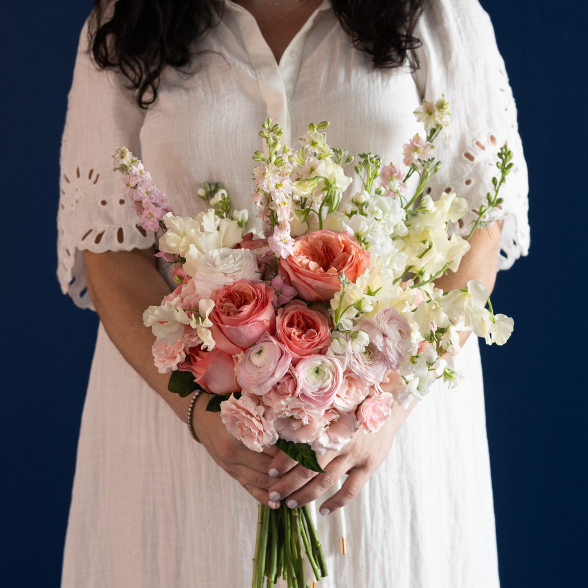 A person in a white eyelet dress holds a large pastel flower bouquet with pink roses, peach blooms, white ranunculus, and green stems against a dark blue background.