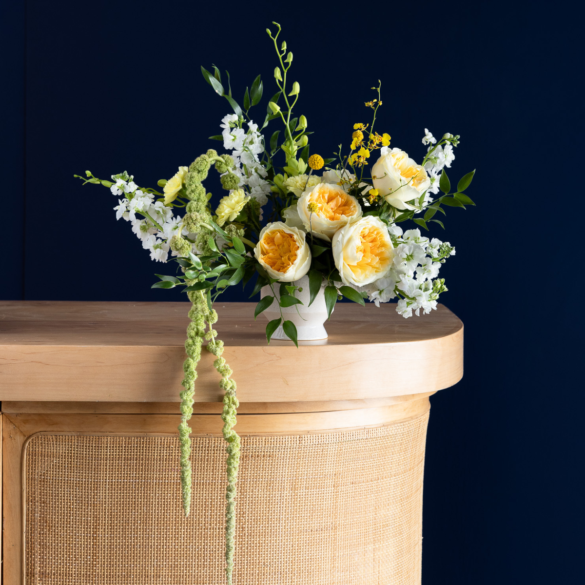 A floral arrangement featuring white and yellow roses, white baby's breath, and green foliage in a small white vase, displayed on a light wood cabinet against a dark navy blue background.