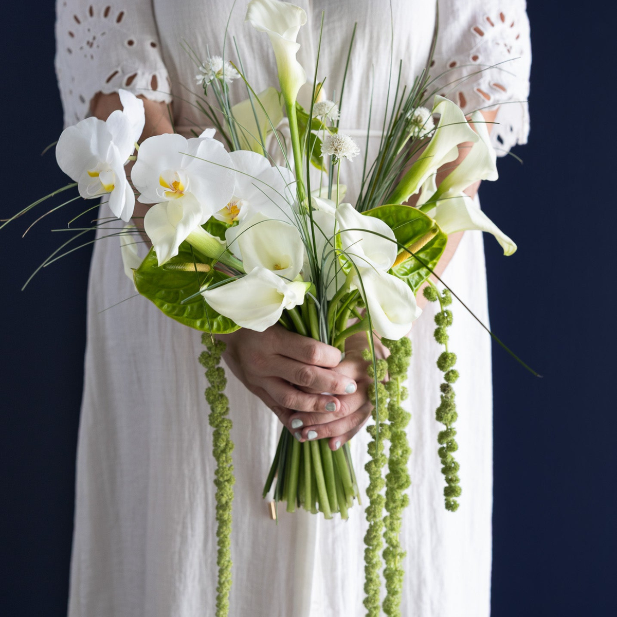 A bouquet of white calla lilies and green foliage held by a person wearing a white eyelet dress against a dark navy background.