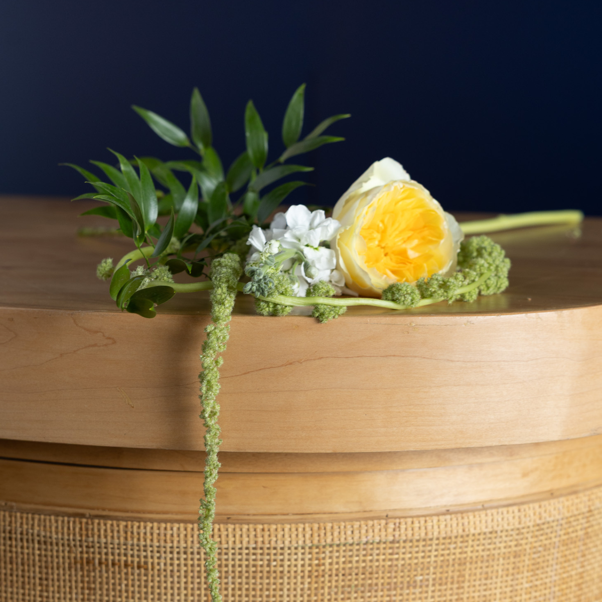 A floral arrangement featuring a yellow-orange tulip, white flowers, and green foliage resting on a light wood surface against a dark navy background.