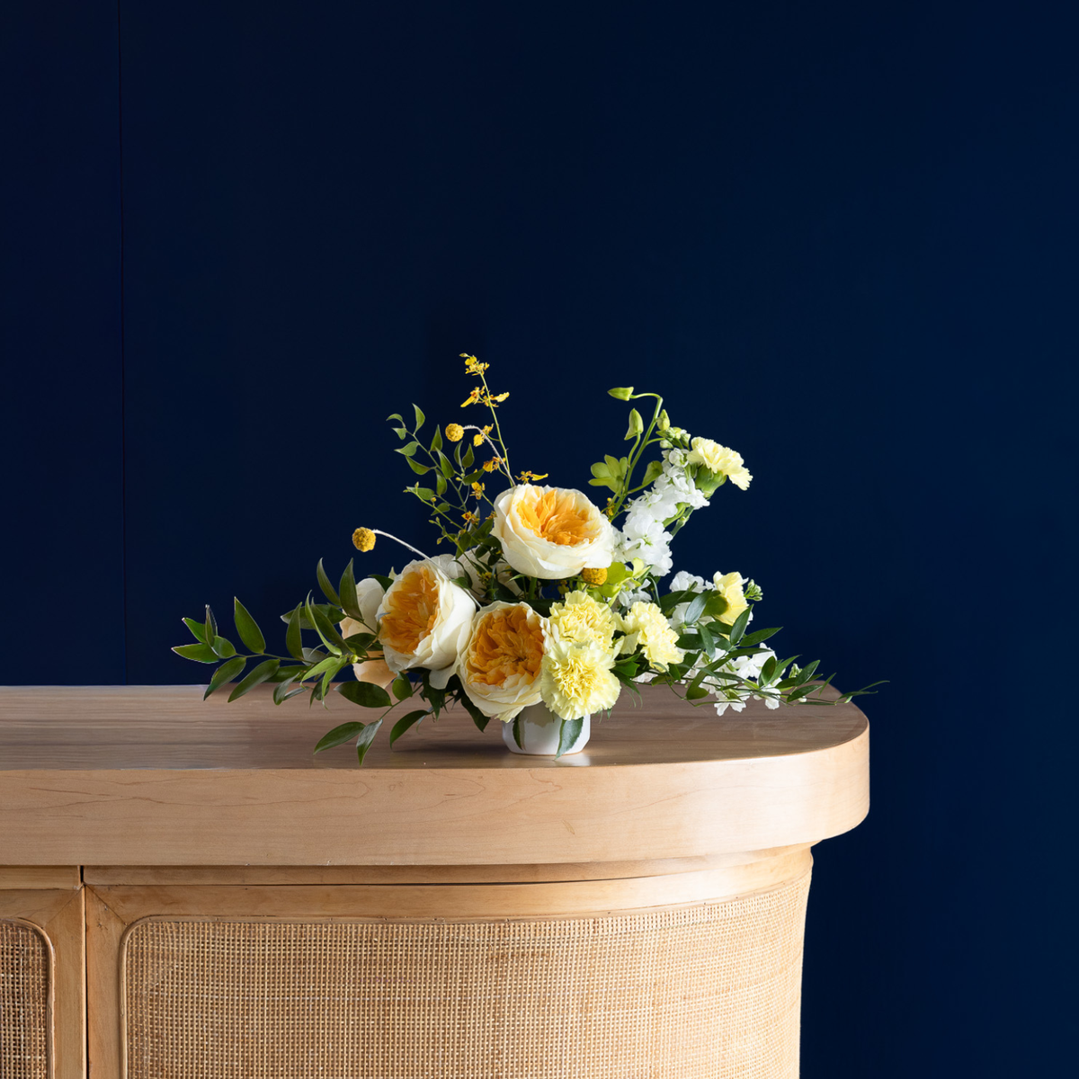 A small floral arrangement with yellow and white flowers and green foliage in a clear vase, placed on a light wood console table against a dark navy blue background.