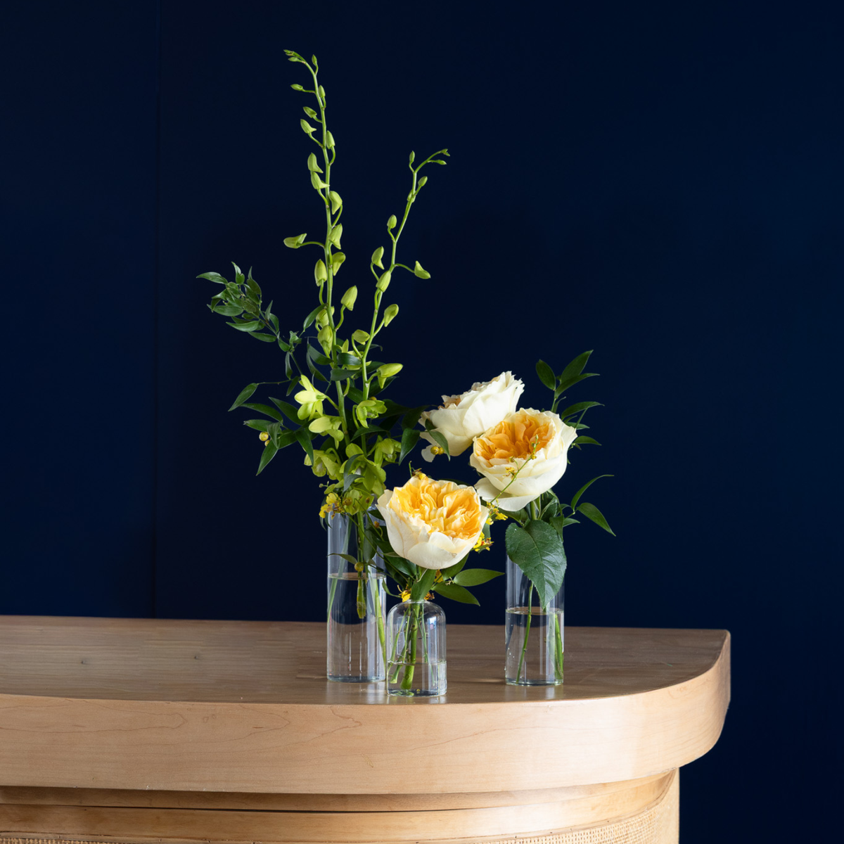 Three clear glass vases holding a floral arrangement with green stems, green leaves, and two pale yellow roses, displayed on a curved light wood console against a dark navy wall.