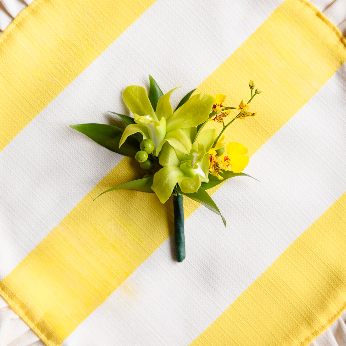 A small floral boutonniere with bright yellow orchids and green leaves, arranged on a white fabric surface with bold yellow diagonal stripes.