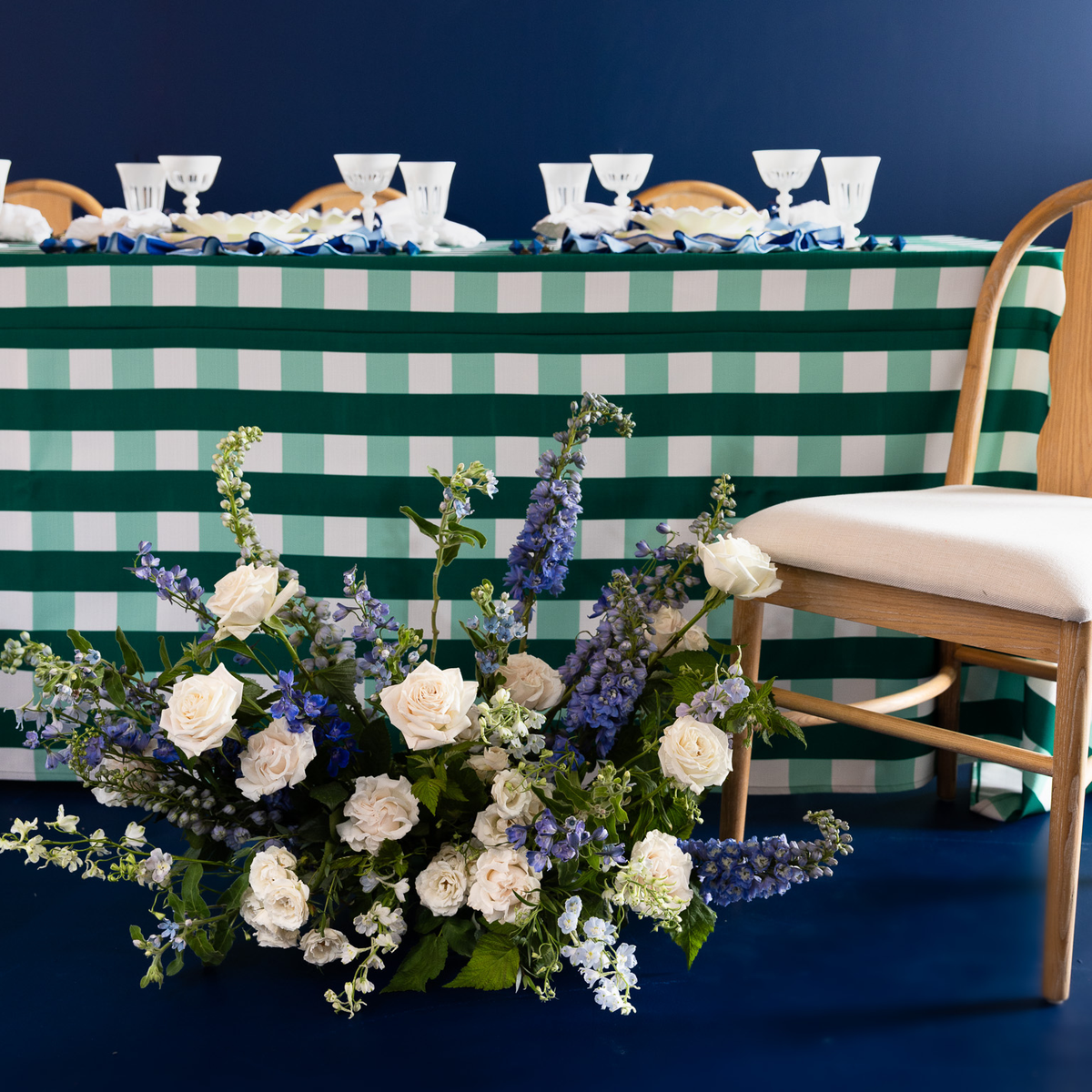 A green and white gingham tablecloth draped over a long table set with white plates and glassware, alongside a wooden chair with a white cushion. In the foreground, a large floral arrangement of white roses, hydrangeas, and blue delphiniums sits on a dark blue floor.