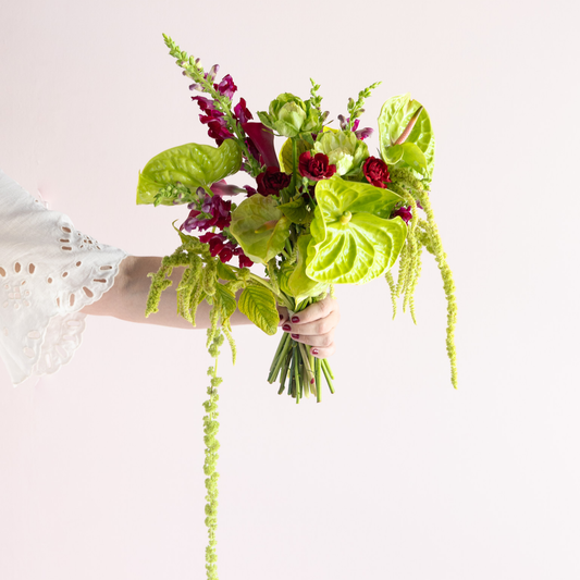 A hand holding a tropical floral bouquet with bright green anthuriums, red roses, and purple flowers against a plain light background.