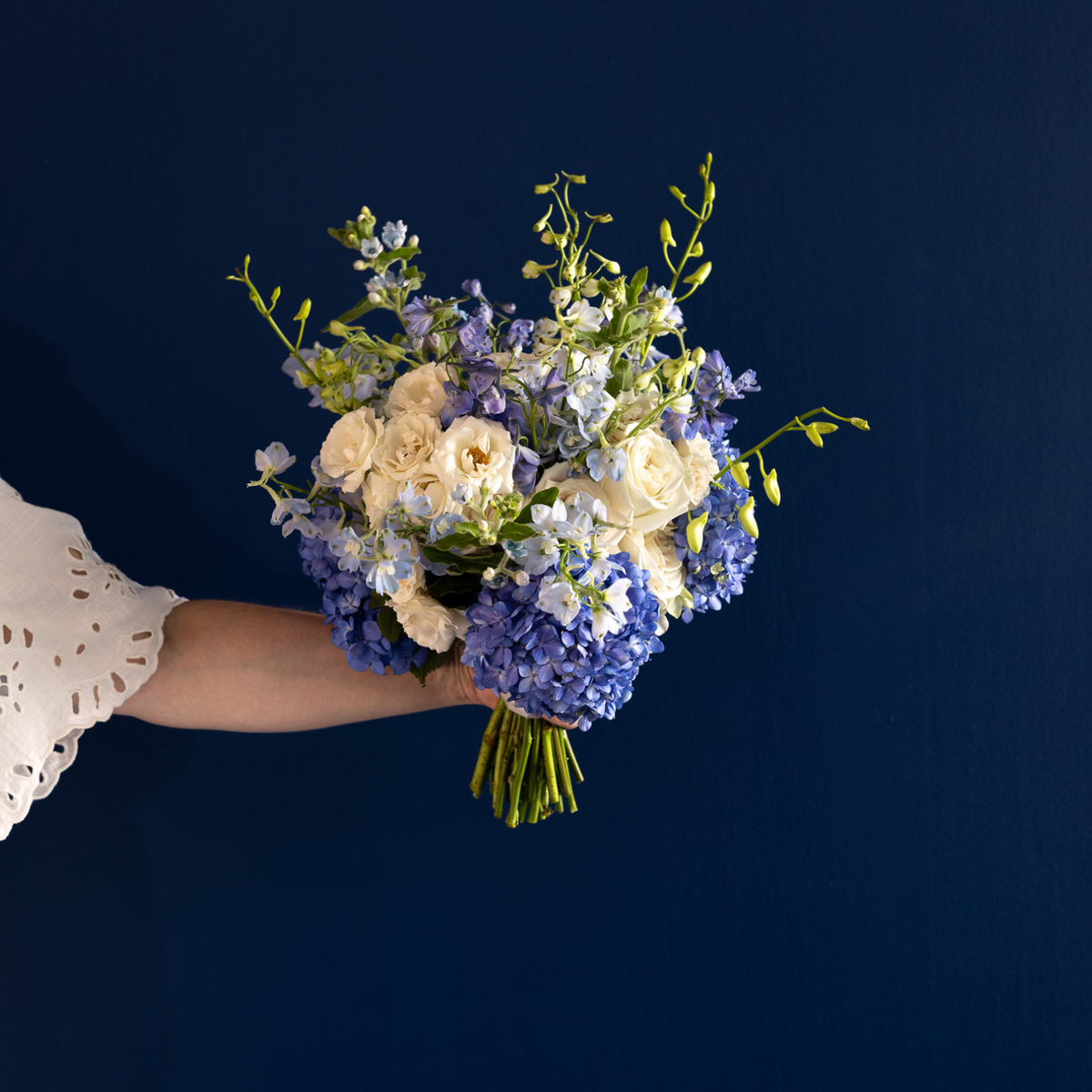 A hand holding a bouquet of blue hydrangeas, lavender delphiniums, cream roses, and green foliage against a solid dark navy blue background.