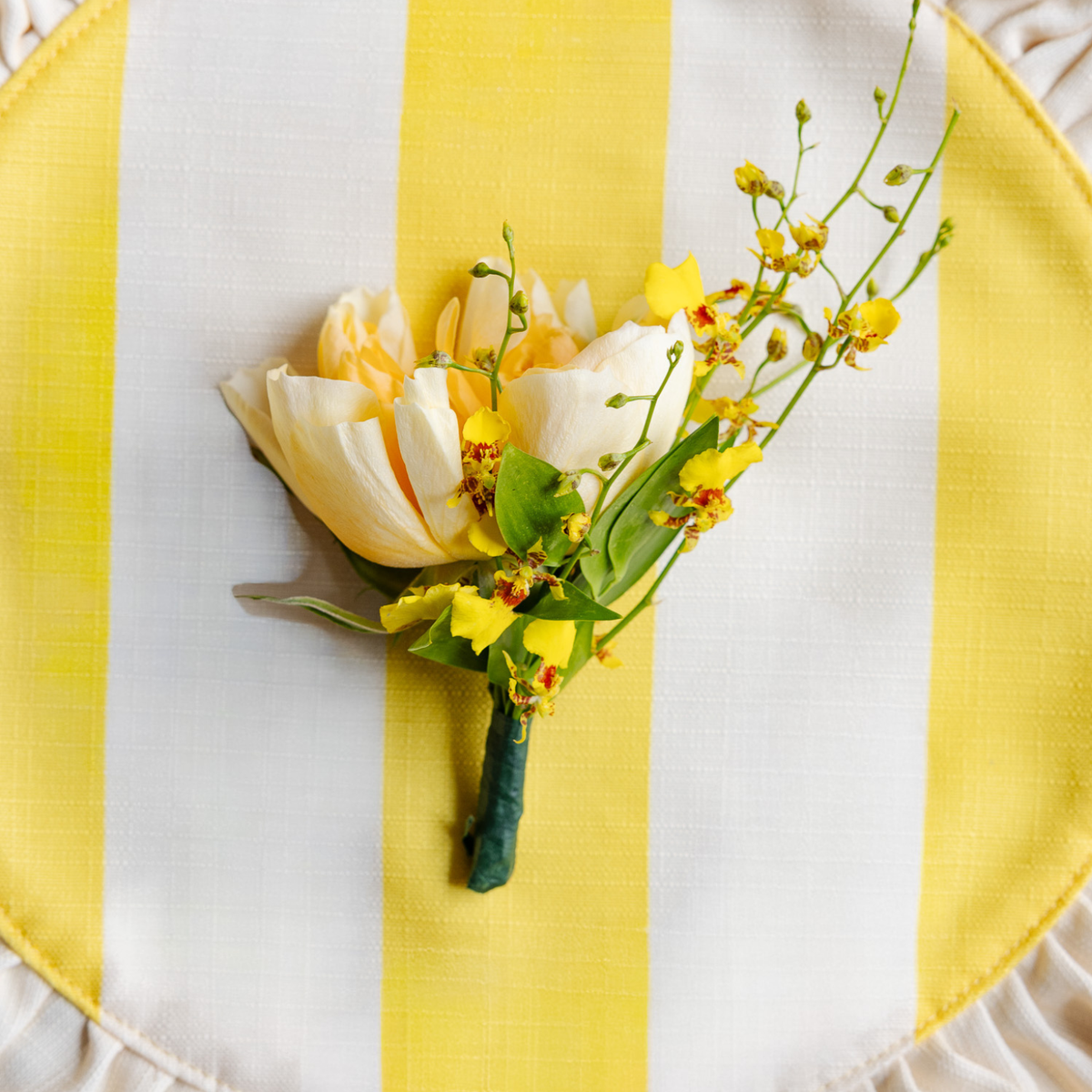 A floral boutonniere featuring a white tulip, yellow orchids, and green leaves, arranged on a yellow and white striped fabric surface.