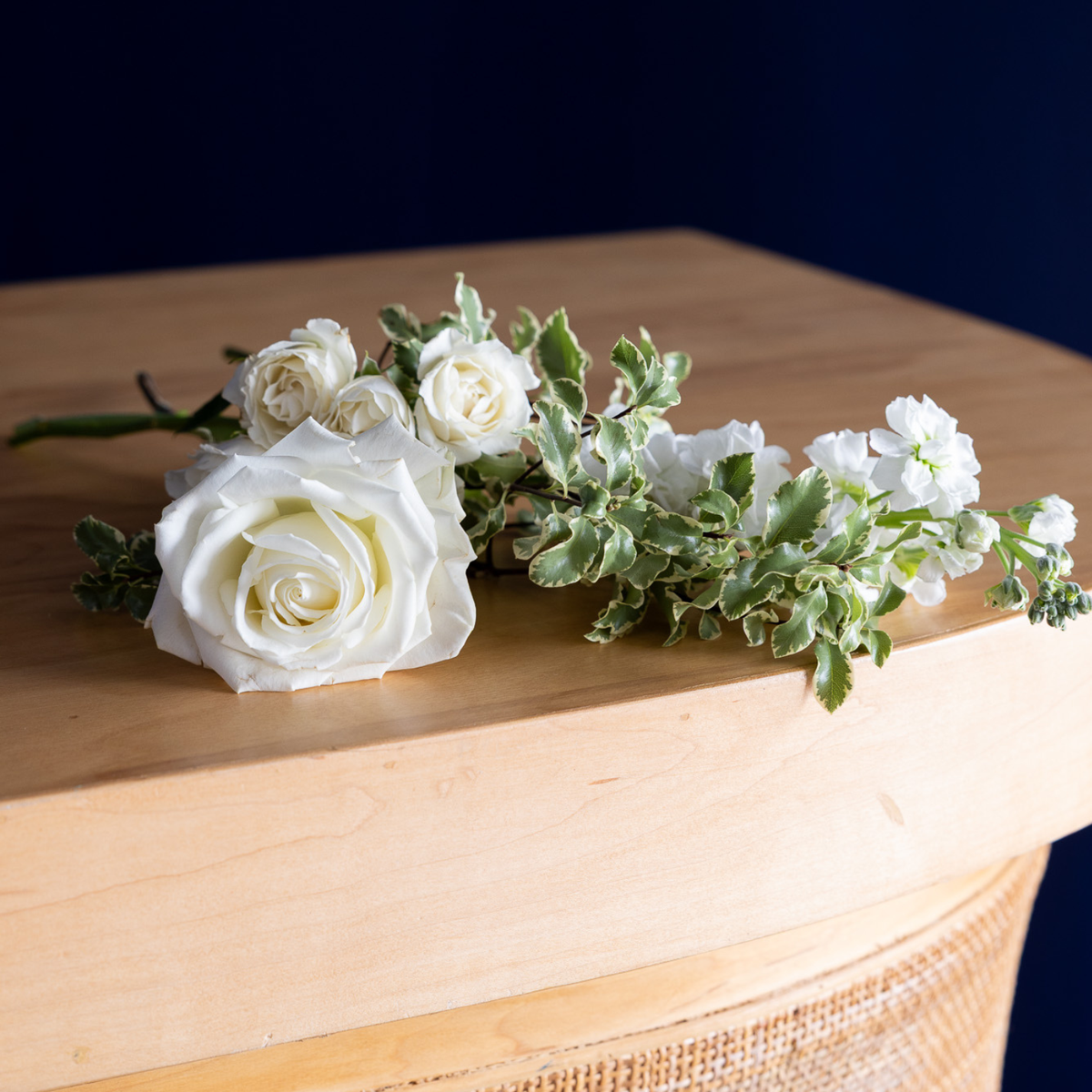 A floral arrangement featuring a large white rose and several small white flowers with variegated green and cream leaves, resting on a light wood surface against a dark navy background.