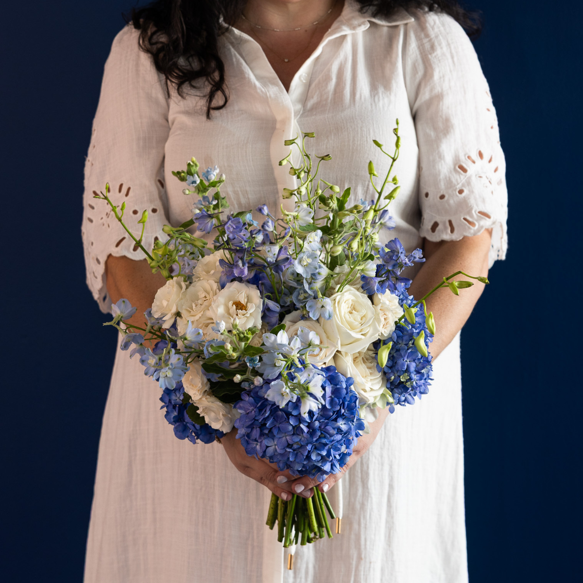 A person in a white short-sleeve dress holds a large bouquet of blue hydrangeas, white roses, and blue delphiniums against a dark navy background.