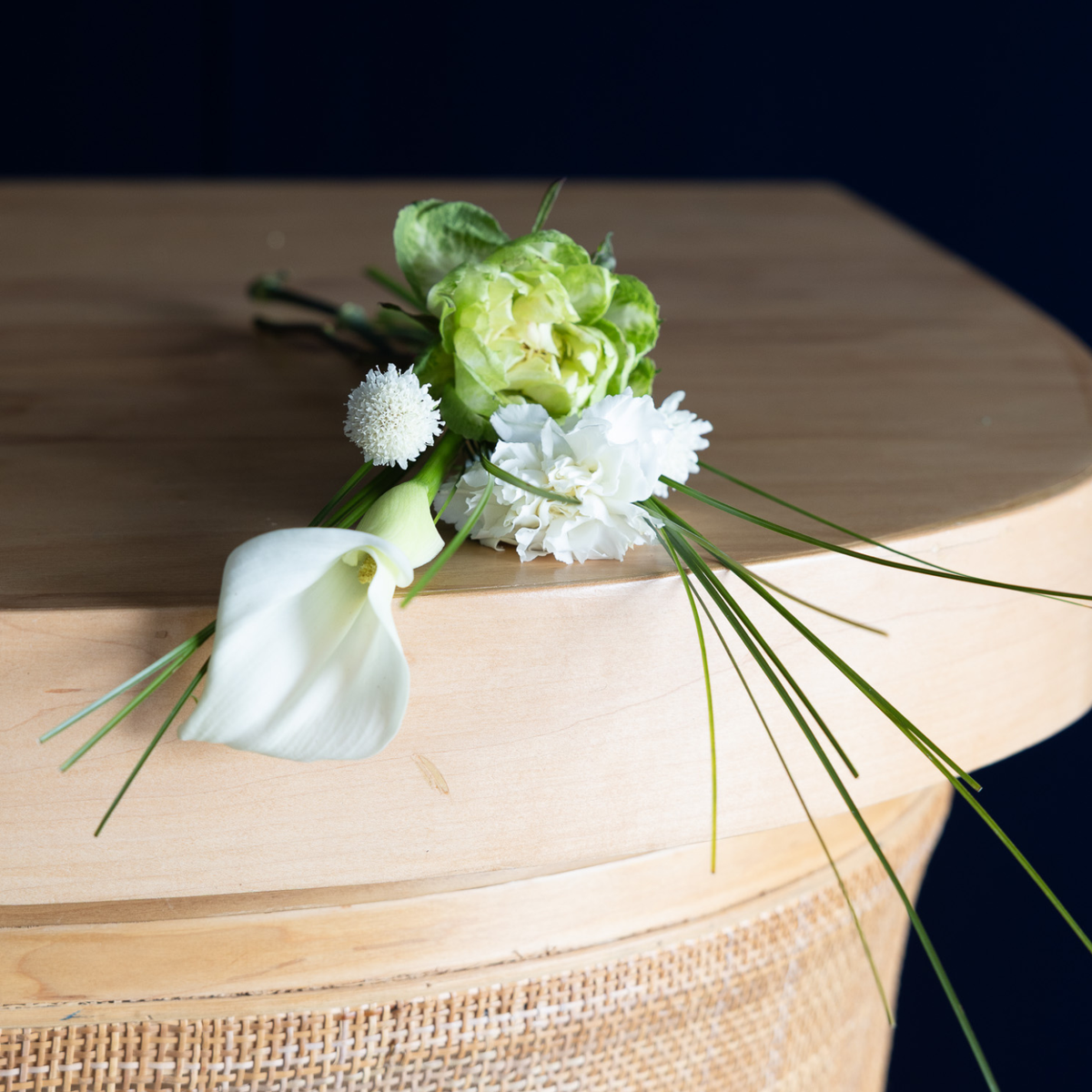 A small floral boutonniere with white and light green flowers and long green stems, resting on a light wood surface against a dark navy background.