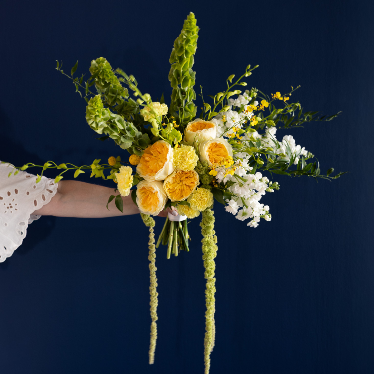 A hand holding a vibrant floral bouquet featuring yellow and orange roses, white daisies, and green foliage against a dark navy blue background.