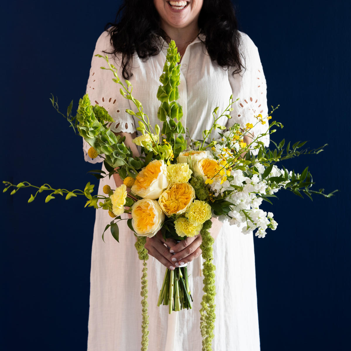 A smiling woman in a white eyelet dress holds a large, vibrant bouquet of yellow and white flowers with green foliage against a dark navy blue background.