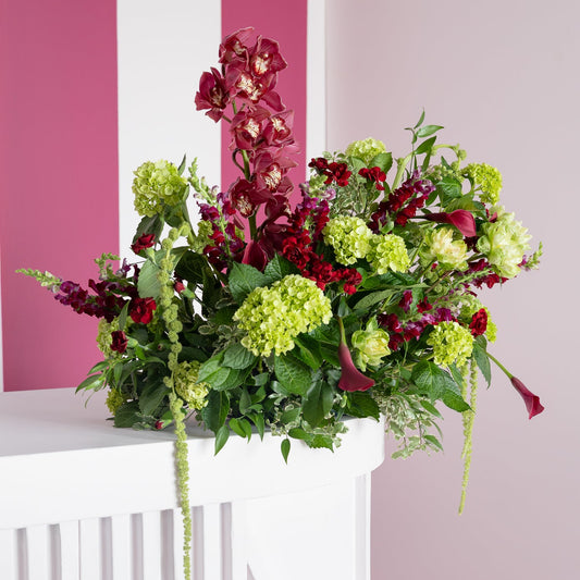 A lush floral arrangement featuring burgundy orchids, light green hydrangea-like blooms, and deep red flowers with green foliage, displayed on a white pedestal against a pink and white striped wall.