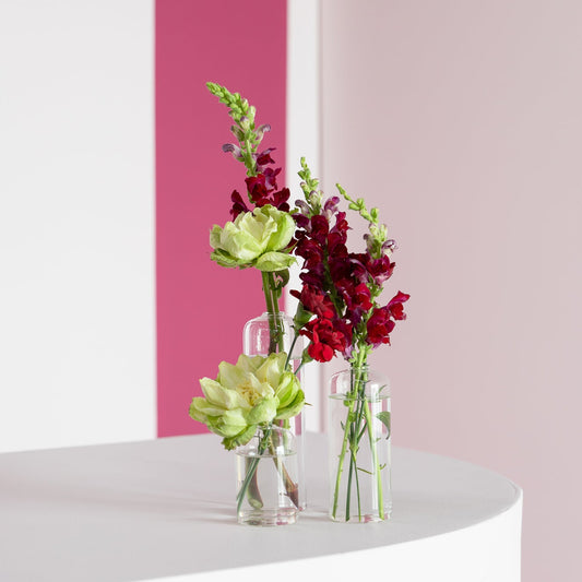 Three clear glass vases with mixed floral arrangements, including pale yellow flowers and deep red blooms, displayed on a white curved table against a pink and white wall.