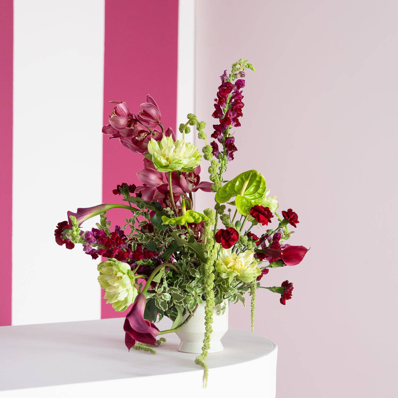 A vibrant artificial flower arrangement in a white ceramic pot, styled on a white wooden ledge against a pink and white striped background.