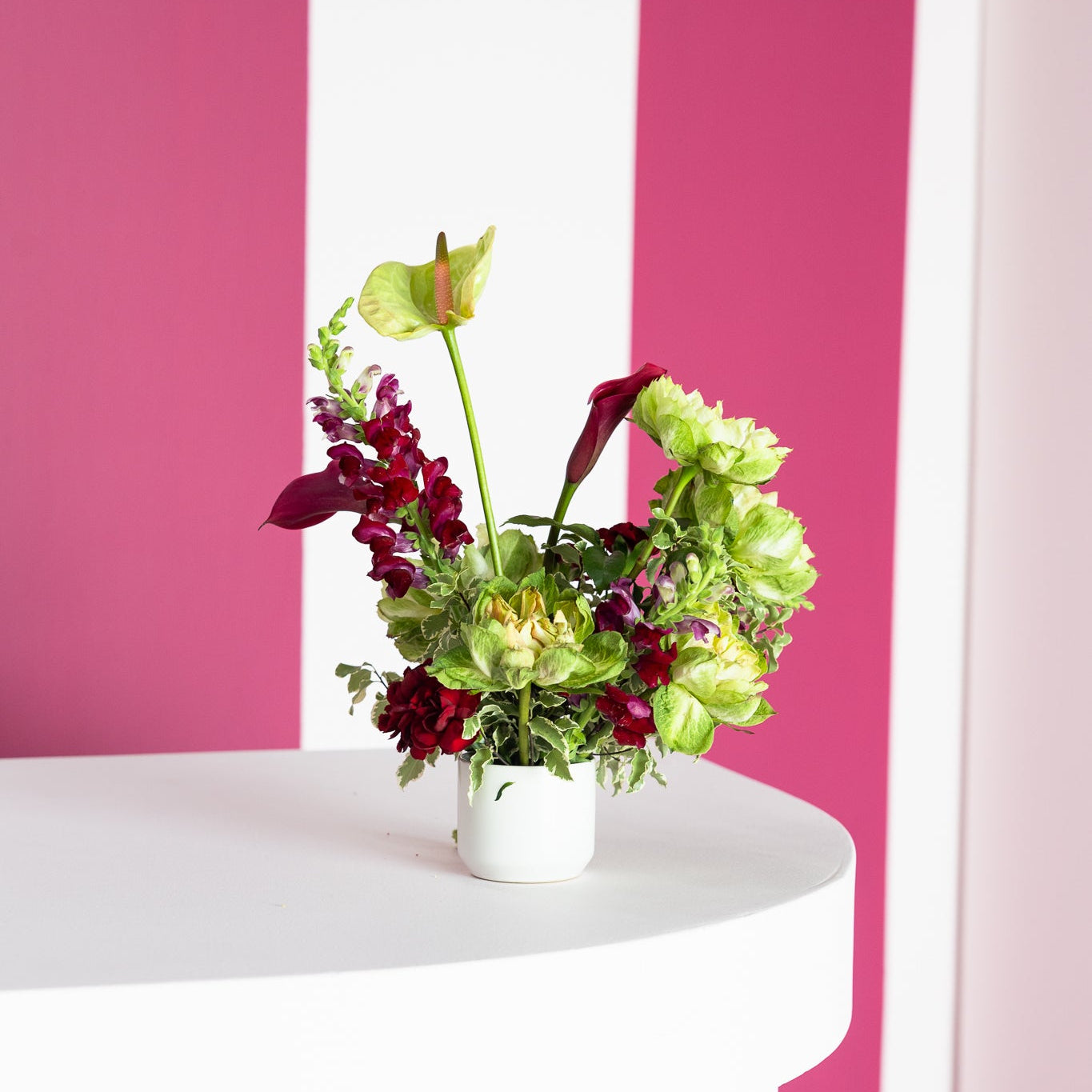 A small white ceramic vase filled with a colorful floral arrangement, placed on a white round table against a bold pink and white striped wall.